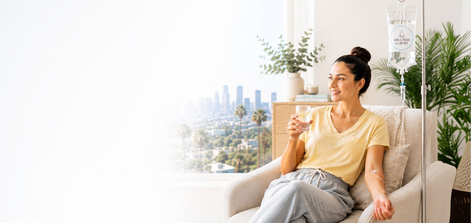 Smiling woman in a yellow t-shirt sipping water during an at-home IV drip with the Los Angeles skyline behind her